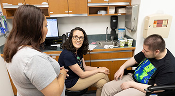 A doctor talks with a male patient in a wheelchair and woman standing nearby.