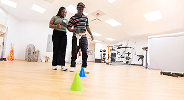 Vanessa Nutakor (DPT student) holding a patient's gait belt while the patient navigates walking around cones in a gym